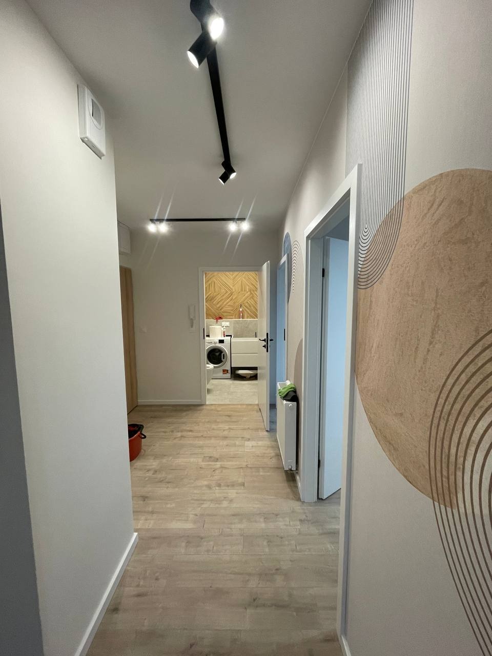 Nowoczesny korytarz z drewnopodobną podłogą, oświetleniem punktowym na suficie i dekoracyjną tapetą z geometrycznym wzorem, widok na łazienkę z pralką.