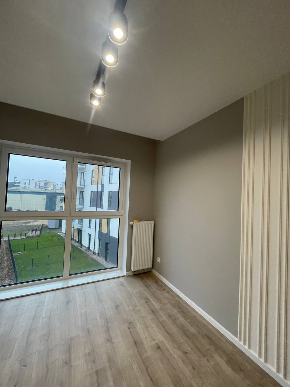 Pusty pokój z panelami ściennymi, oknem z widokiem na osiedle i lampą sufitową z pięcioma reflektorami.