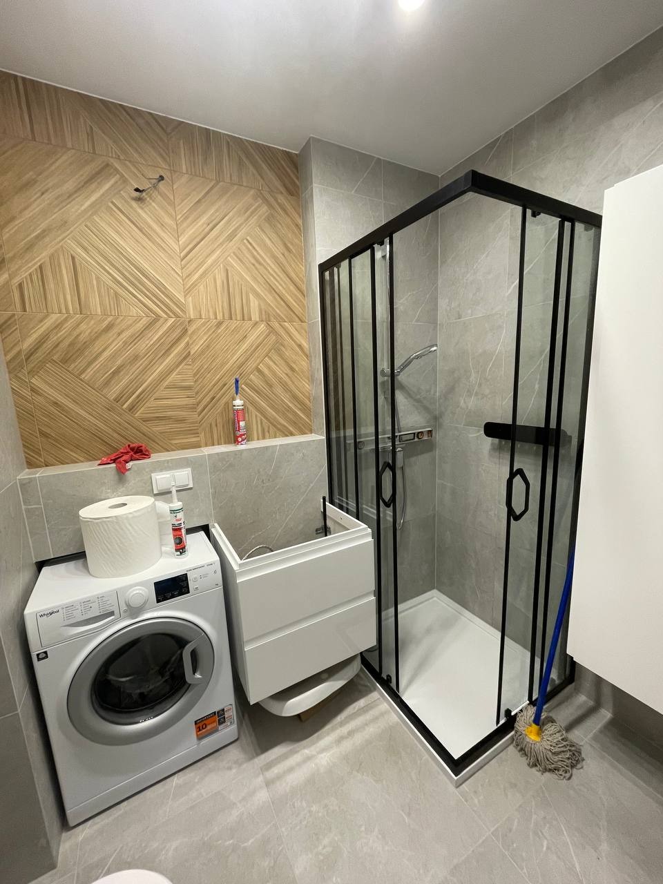 Nowoczesna łazienka z kabiną prysznicową w czarnej ramie, pralką, umywalką z szafką i mozaiką drewnopodobną na ścianie. Na pralce leżą ręczniki papierowe i silikon.