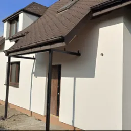 Jasna elewacja domu jednorodzinnego z brązowym dachem i stolarką okienną, widok z boku na tle zieleni. Nowoczesne wykończenie, boczny widok.