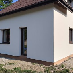 Złota rączka - Świeżo otynkowany, kremowy dom jednorodzinny z brązowym dachem i ramami okiennymi, otoczony zielenią i kamienistym podłożem, w słoneczny dzień.