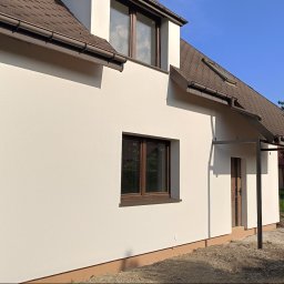 Złota rączka - Jasny, otynkowany dom jednorodzinny z brązowym dachem i stolarką okienną. Widoczne detale elewacji, błękitne niebo i fragment zieleni w tle. Nowoczesna architektura.