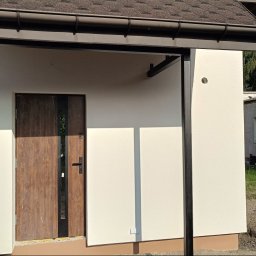 Złota rączka - Otynkowana elewacja budynku z brązowymi elementami wykończeniowymi, widoczna boczna ściana z drzwiami wejściowymi i fragment dachu z rynną.