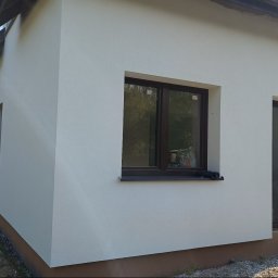 Złota rączka - Biała elewacja budynku z brązowymi oknami i podmurówką. Widoczna struktura tynku i detale wykończenia. Budynek oświetlony naturalnym światłem słonecznym.