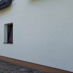 Złota rączka - Ściana budynku po ociepleniu i malowaniu, widoczne okna z brązowymi ramami i biała elewacja z delikatną fakturą. Na dachu zwisają kable.