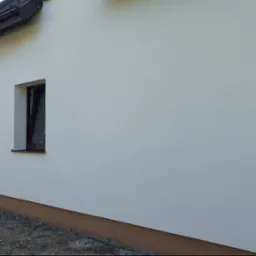 Ściana budynku po ociepleniu i malowaniu, widoczne okna z brązowymi ramami i biała elewacja z delikatną fakturą. Na dachu zwisają kable.