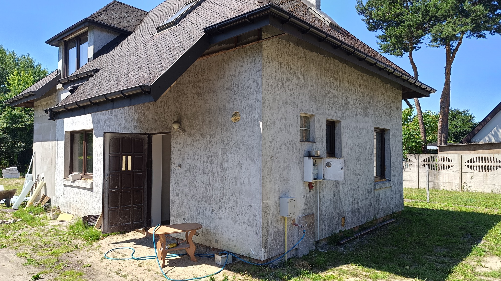 Szara elewacja domu jednorodzinnego z widocznymi elementami instalacji zewnętrznej, stolarką okienną i drzwiami wejściowymi. W tle zieleń drzew.