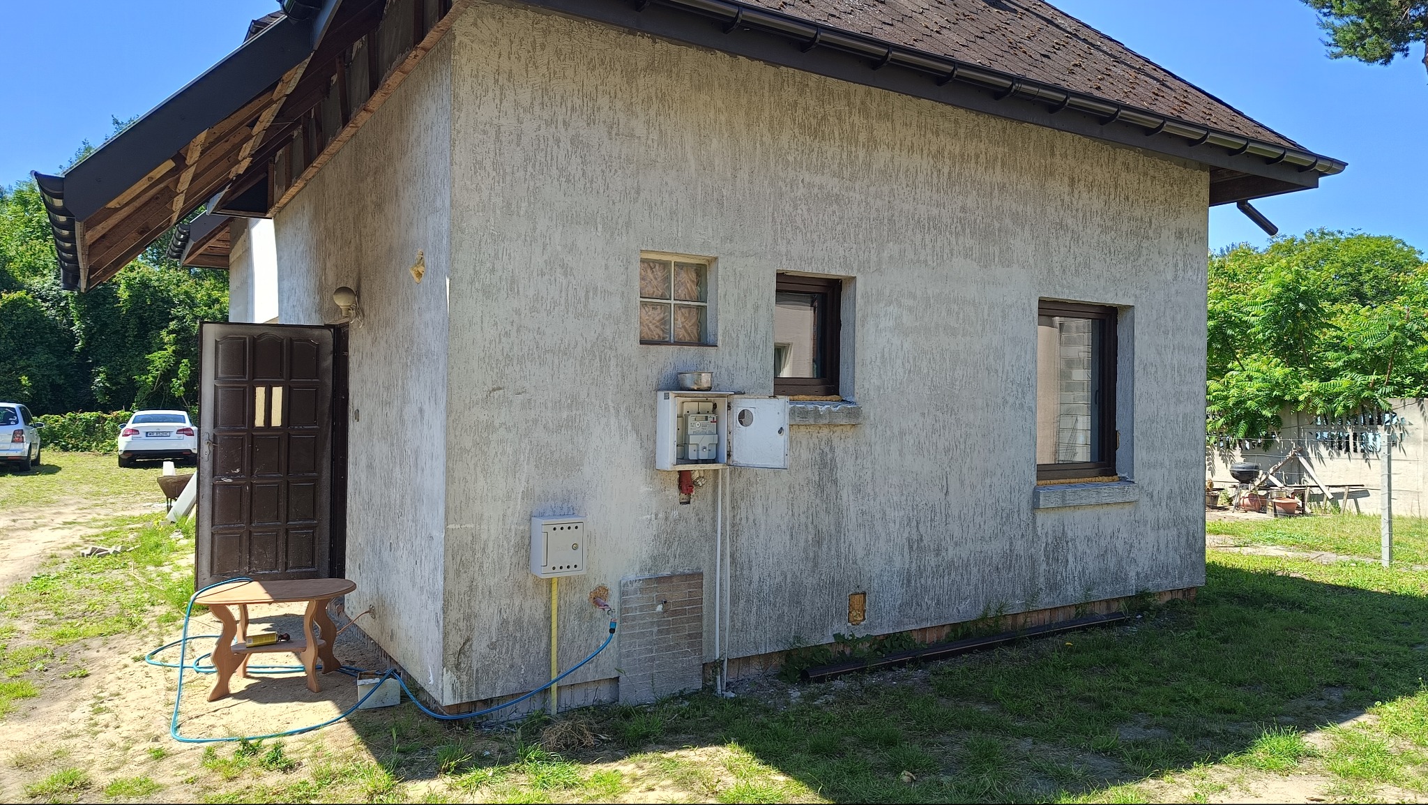 Szara elewacja budynku z fakturą baranka, widoczne okna, drzwi z brązowego drewna i przyłącza. W tle zieleń i samochody. Widoczny fragment dachu.