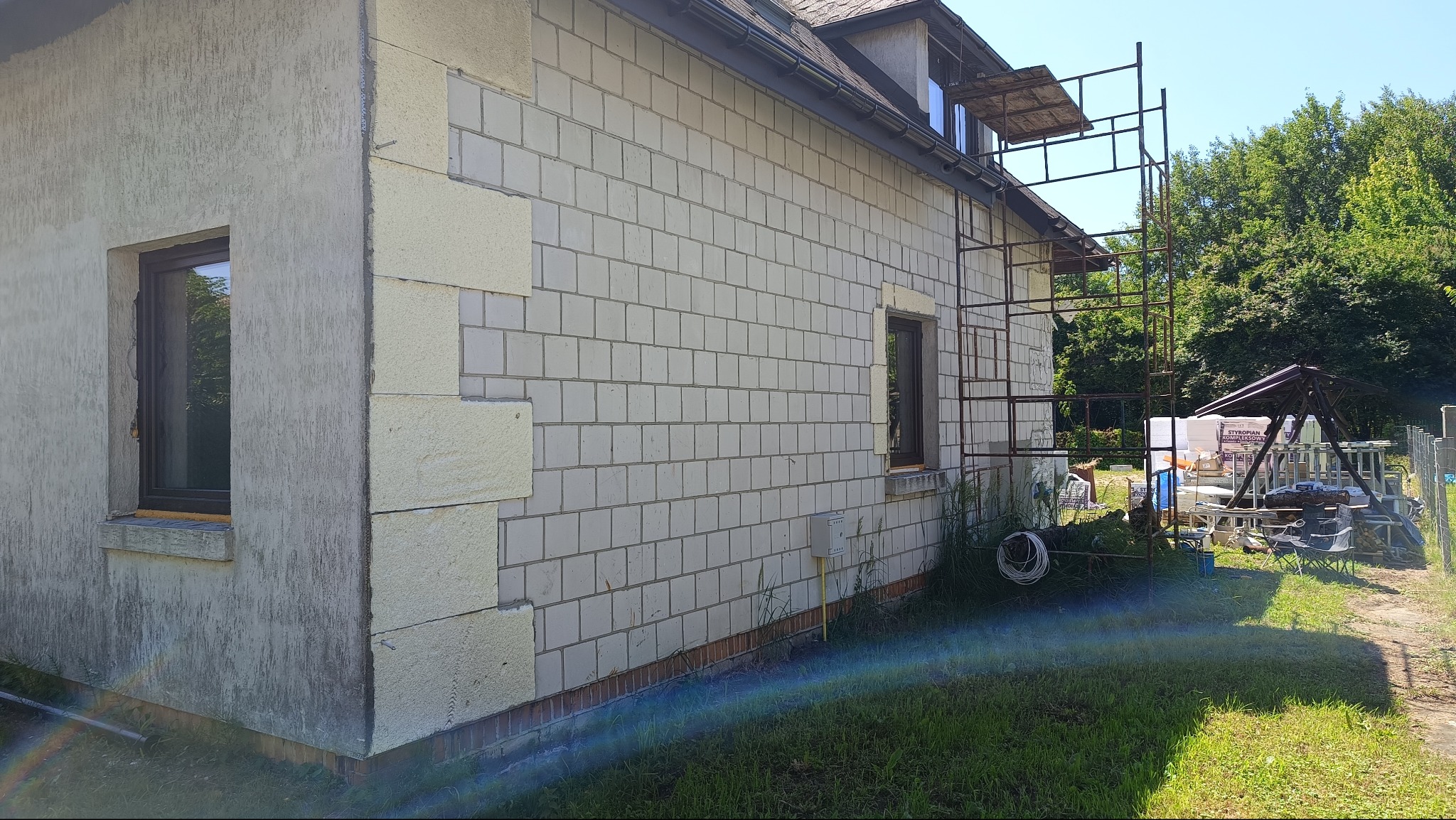 Ściana domu z częściowo ocieploną elewacją, widoczne rusztowanie i przygotowane materiały. Fragment elewacji jest otynkowany, a część w trakcie ocieplania.