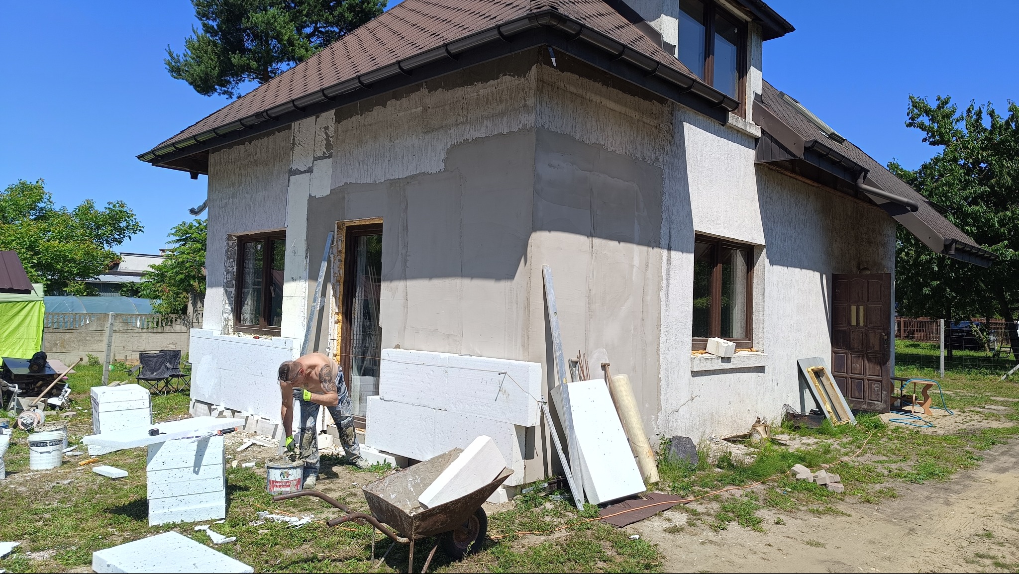 Prace ociepleniowe na elewacji domu jednorodzinnego. Widoczne płyty styropianowe, częściowo otynkowana ściana, pracownik w trakcie nakładania zaprawy. Kątowe ujęcie z zewnątrz.