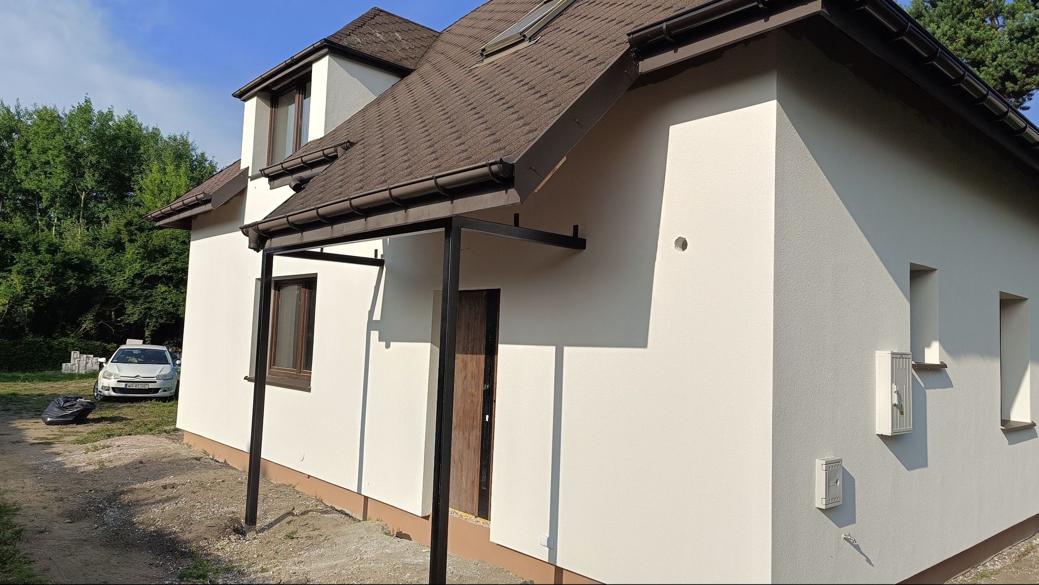 Jasna elewacja domu jednorodzinnego z brązowym dachem i stolarką okienną, widok z boku na tle zieleni. Nowoczesne wykończenie, boczny widok.