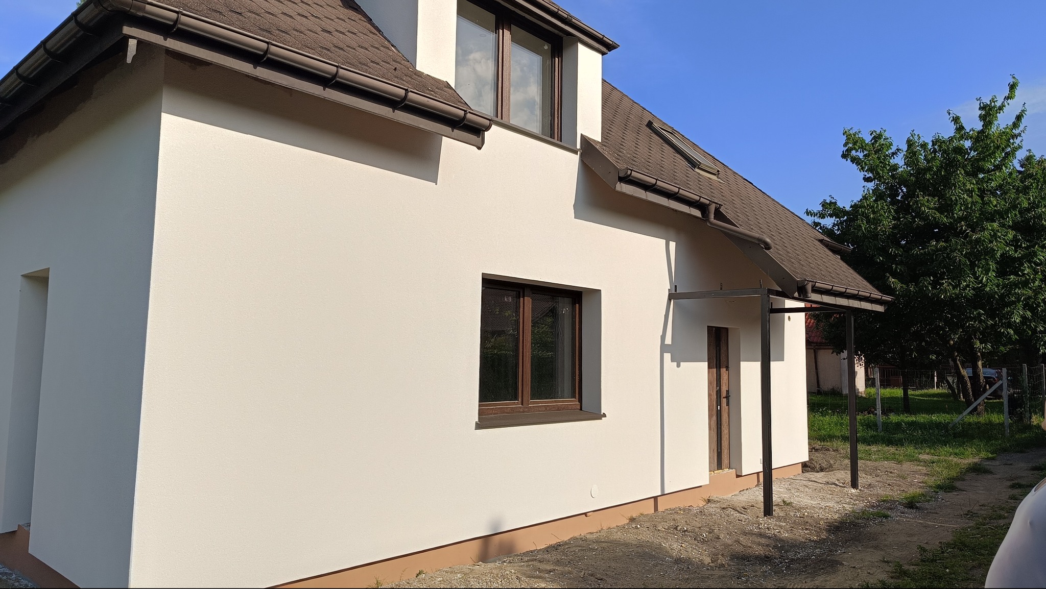 Jasny, otynkowany dom jednorodzinny z brązowym dachem i stolarką okienną. Widoczne detale elewacji, błękitne niebo i fragment zieleni w tle. Nowoczesna architektura.