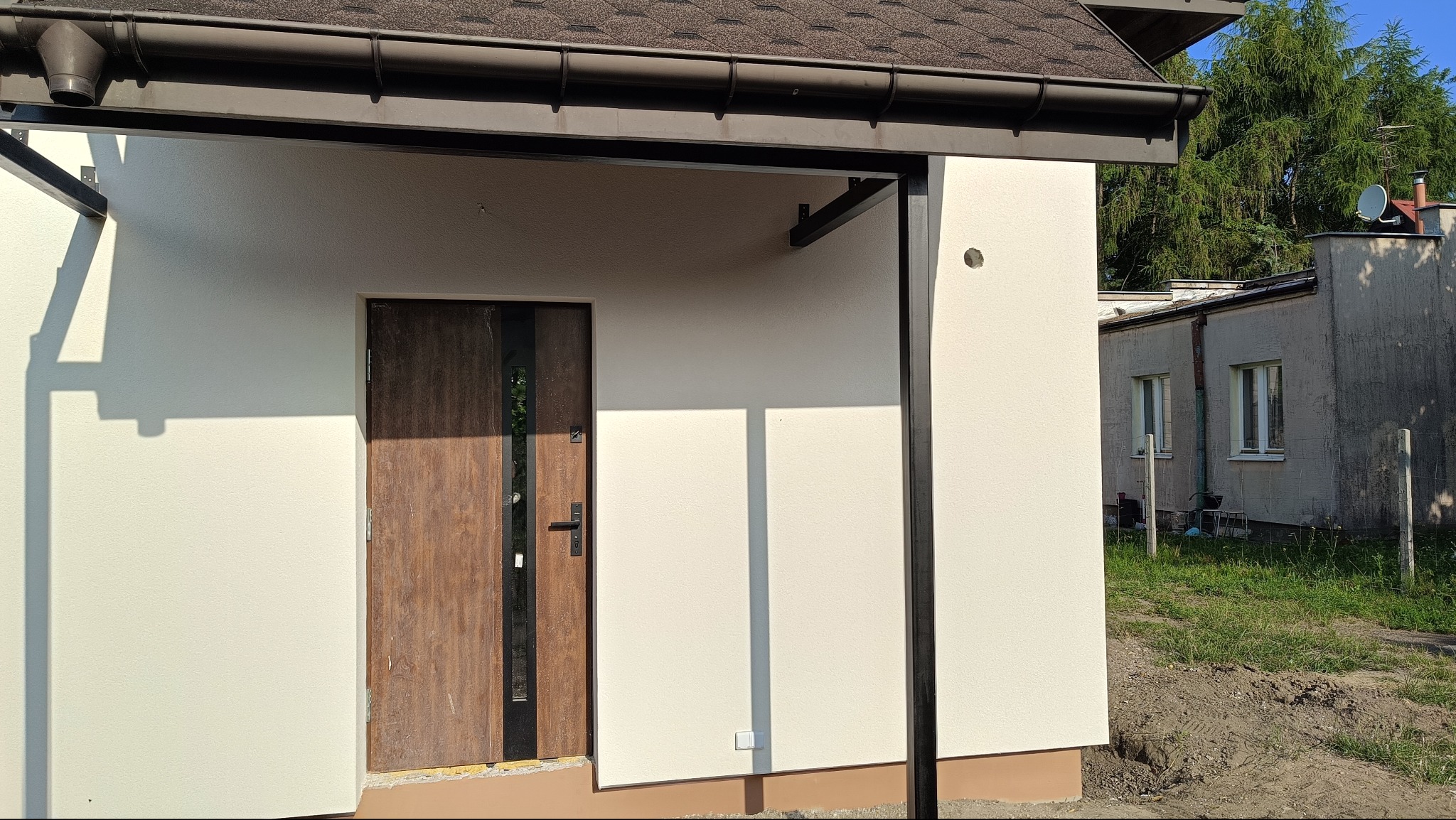 Otynkowana elewacja budynku z brązowymi elementami wykończeniowymi, widoczna boczna ściana z drzwiami wejściowymi i fragment dachu z rynną.