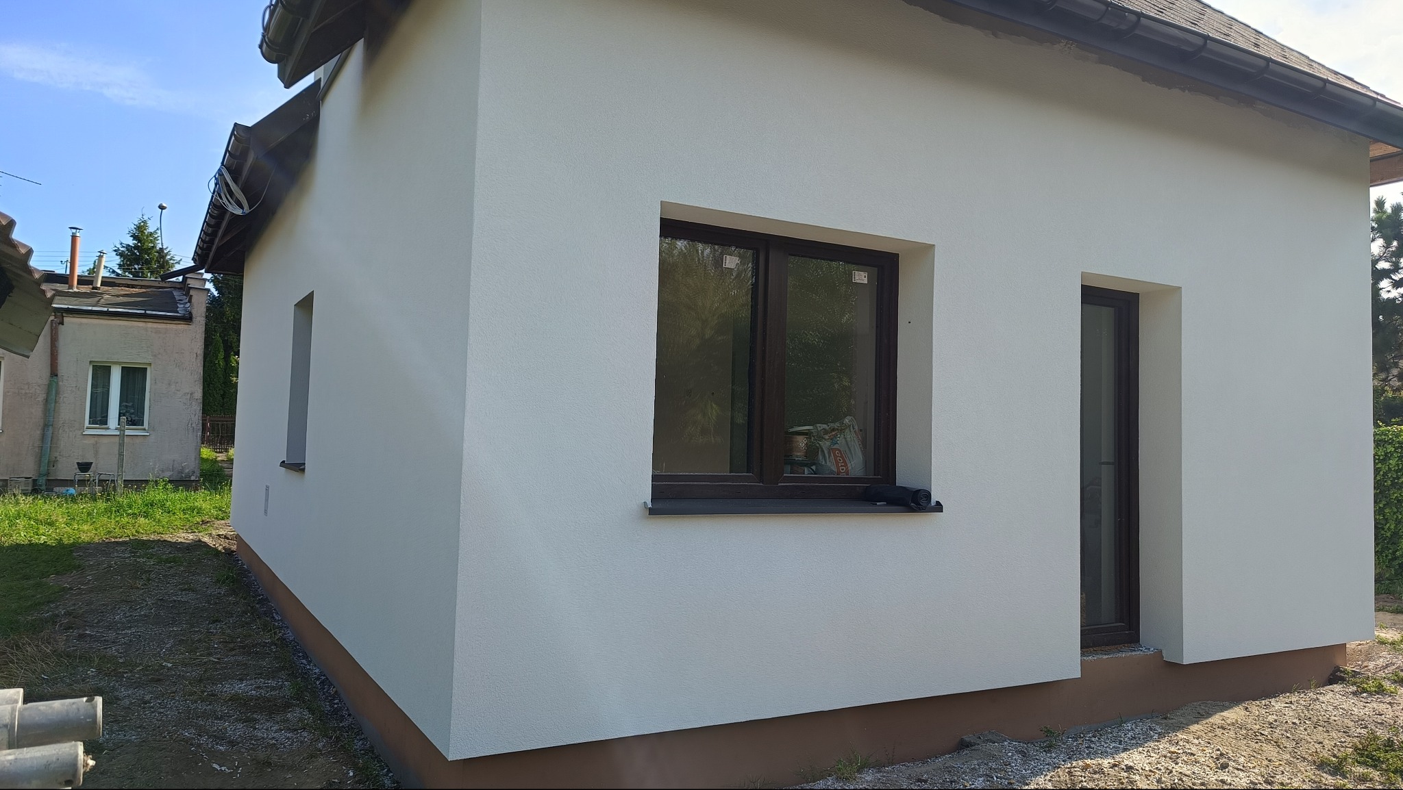 Biała elewacja budynku z brązowymi oknami i podmurówką. Widoczna struktura tynku i detale wykończenia. Budynek oświetlony naturalnym światłem słonecznym.