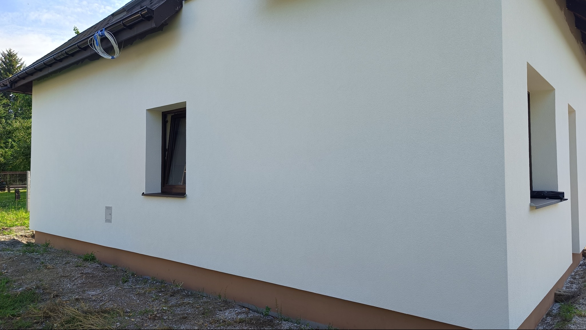 Ściana budynku po ociepleniu i malowaniu, widoczne okna z brązowymi ramami i biała elewacja z delikatną fakturą. Na dachu zwisają kable.
