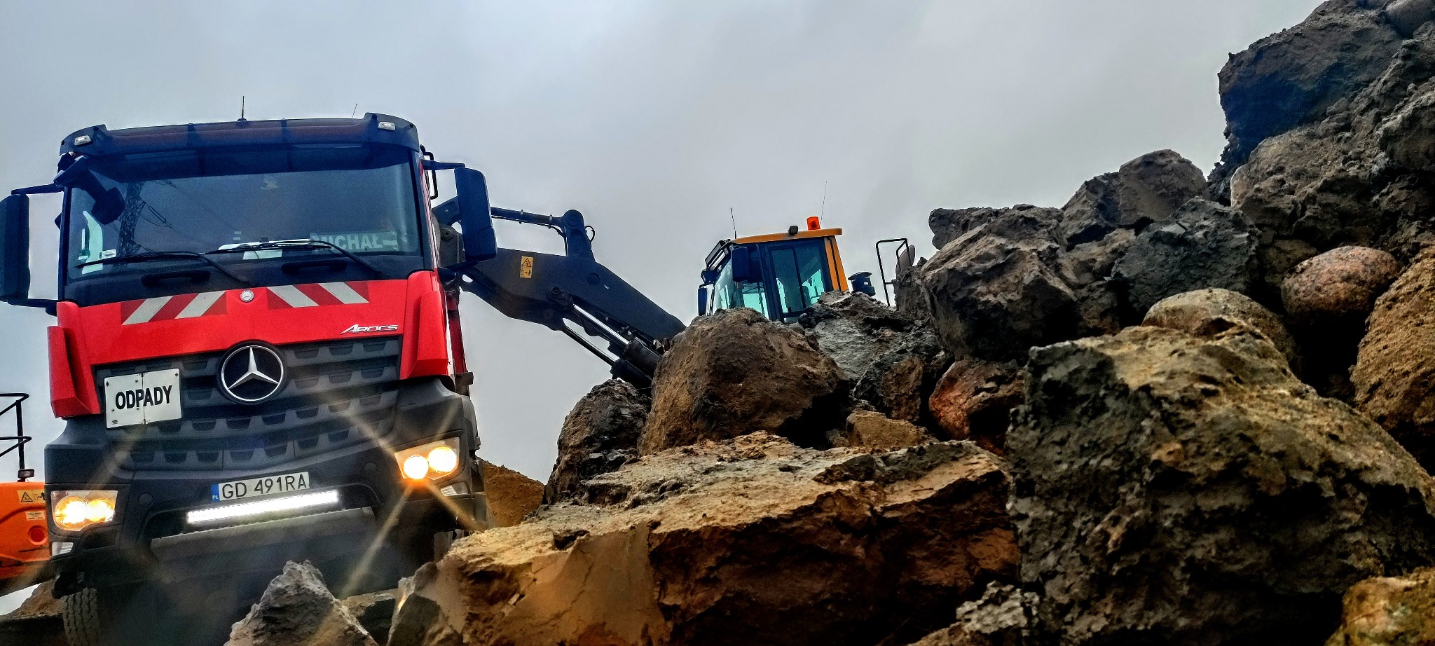Czerwona ciężarówka z napisem ODPADY i żółta koparka na tle sterty dużych kamieni i gruzu pod pochmurnym niebem.