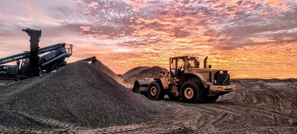 Żółta ładowarka Volvo na tle zachodzącego słońca, obok hałda kruszywa i kruszarka, widoczne ślady opon na ziemi.