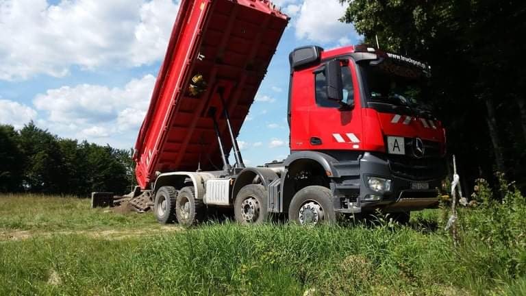Czerwona wywrotka Mercedes z podniesioną skrzynią ładunkową, stojąca na łące z wysoką trawą, z wysypanym niewielkim kopcem ziemi. W tle widoczny las i błękitne niebo z chmurami.