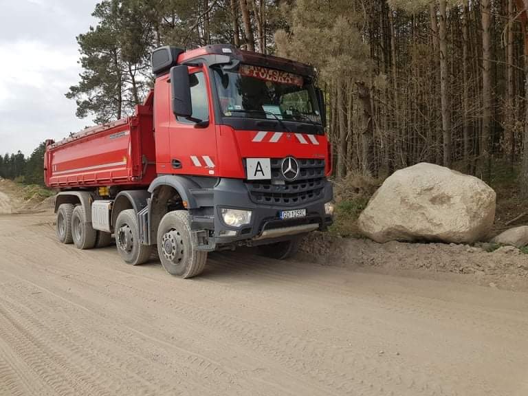 Czerwona wywrotka Mercedes-Benz z napisem 'Polska' na przedniej szybie, zaparkowana na piaszczystej drodze obok dużego, jasnego kamienia polnego na tle lasu sosnowego.