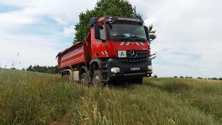Czerwona wywrotka Mercedes-Benz z plandeką, zaparkowana na zarośniętym polu z widocznym drzewem w tle i błękitnym niebem z chmurami.