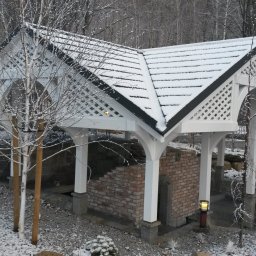 MARGAB STOLARSTWO OGÓLNE MARCIN CEREMON - Zadaszona konstrukcja z białego drewna z kratownicą i czarnym dachem pokrytym śniegiem, wsparta na białych kolumnach z ceglaną podstawą, otoczona zimowym krajobrazem z drzewami i śniegiem.