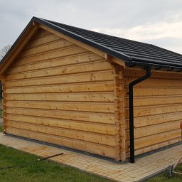 MARGAB STOLARSTWO OGÓLNE MARCIN CEREMON - Zewnętrzny widok na mały budynek z bali o jasnym drewnie, z czarnym dachem i rynnami, osadzony na płytkach chodnikowych.