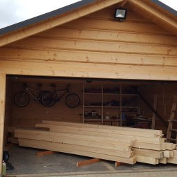 MARGAB STOLARSTWO OGÓLNE MARCIN CEREMON - Wnętrze drewnianej konstrukcji z widocznymi belkami, rowerami zawieszonymi na ścianie, półkami z narzędziami w tle i stosem obrobionego drewna na pierwszym planie.