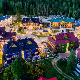 MARGAB STOLARSTWO OGÓLNE MARCIN CEREMON - Panoramiczny widok z lotu ptaka na kompleks budynków mieszkalnych o zróżnicowanej architekturze, otoczonych lasem i oświetlonych dekoracyjnymi lampami.