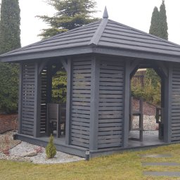 MARGAB STOLARSTWO OGÓLNE MARCIN CEREMON - Drewniana altana pomalowana na szaro, z meblami wewnątrz, otoczona zielonym trawnikiem, ozdobnymi krzewami i kamieniami.