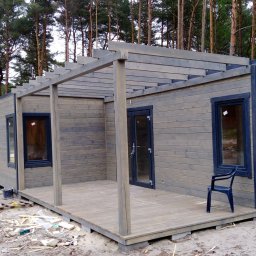 MARGAB STOLARSTWO OGÓLNE MARCIN CEREMON - Szereg drewnianych domków letniskowych w kolorze szaro-brązowym, zadaszone drewnianą pergolą, w trakcie budowy, widoczne elementy konstrukcyjne i śmieci budowlane na piaszczystym podłożu, w tle...