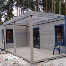 Szereg drewnianych domków letniskowych w kolorze szaro-brązowym, zadaszone drewnianą pergolą, w trakcie budowy, widoczne elementy konstrukcyjne i śmieci budowlane na piaszczystym podłożu, w tle...