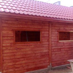 MARGAB STOLARSTWO OGÓLNE MARCIN CEREMON - Czerwona drewniana konstrukcja w trakcie malowania, widoczne okna bez szyb, blaszany dach i mężczyzna z pędzlem.