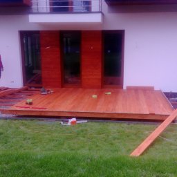 MARGAB STOLARSTWO OGÓLNE MARCIN CEREMON - Budowa drewnianego tarasu przy nowoczesnym domu z drewnianą elewacją, widoczni dwaj pracownicy, jeden w czerwonej kurtce stoi na konstrukcji, drugi pracuje na ziemi, narzędzia rozłożone na trawie.