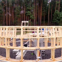 MARGAB STOLARSTWO OGÓLNE MARCIN CEREMON - Konstrukcja szkieletowa budynku w trakcie budowy, widoczna na tle lasu, z detalami takimi jak drabina, bloczki betonowe i czarne kamienie na ziemi.