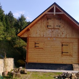 MARGAB STOLARSTWO OGÓLNE MARCIN CEREMON - Drewniany domek z okiennicami, wykończony ciemną podmurówką i ozdobną listwą pod dachem, otoczony zielenią i kamiennym murkiem.