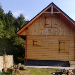 Drewniany domek z okiennicami, wykończony ciemną podmurówką i ozdobną listwą pod dachem, otoczony zielenią i kamiennym murkiem.