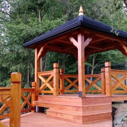 MARGAB STOLARSTWO OGÓLNE MARCIN CEREMON - Drewniana altana z czarnym dachem, otoczona zielenią drzew, z barierkami w kształcie krzyżyków i schodkami prowadzącymi na podest.