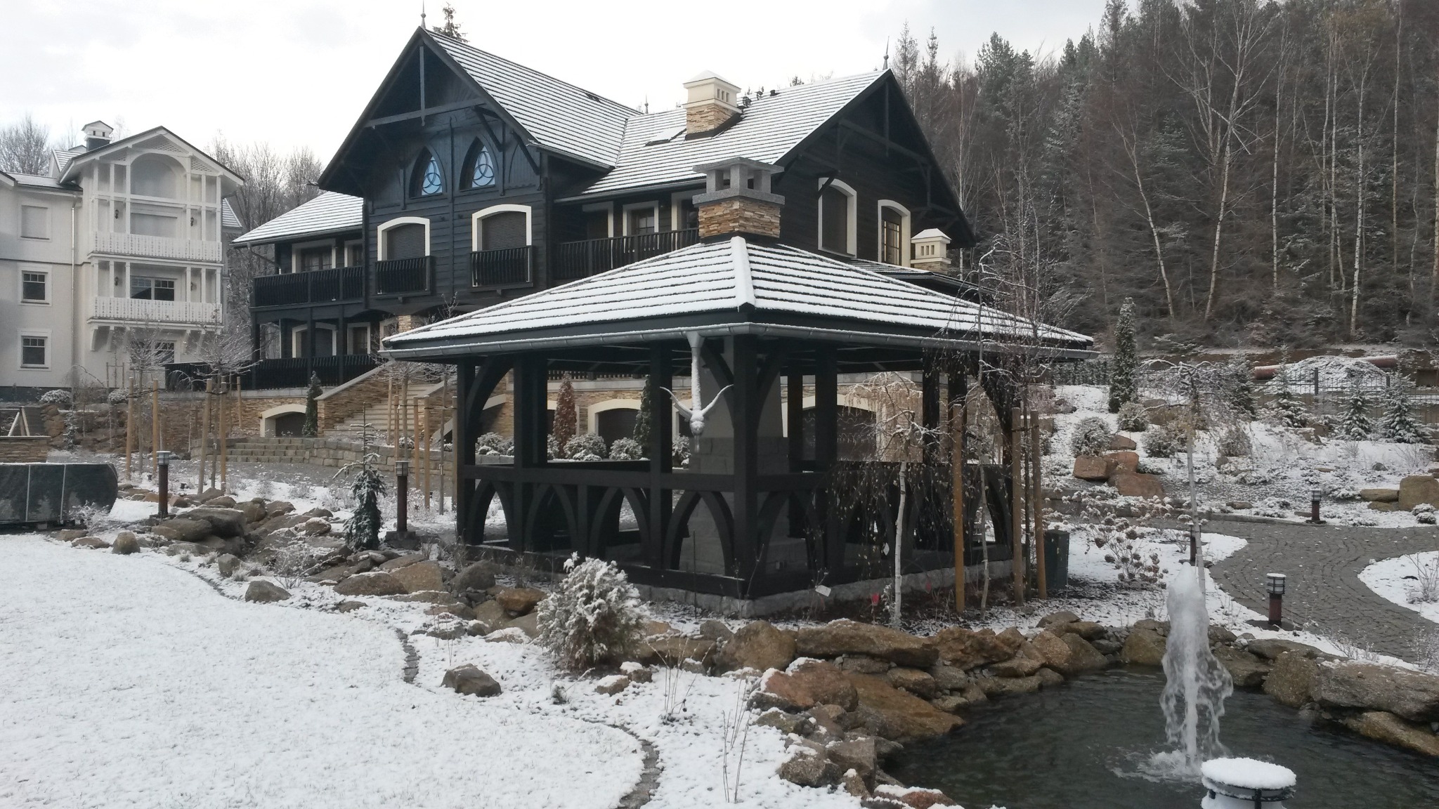 Czarna altana z rzeźbą ptaka na tle domu w stylu góralskim i ośnieżonego ogrodu z fontanną.