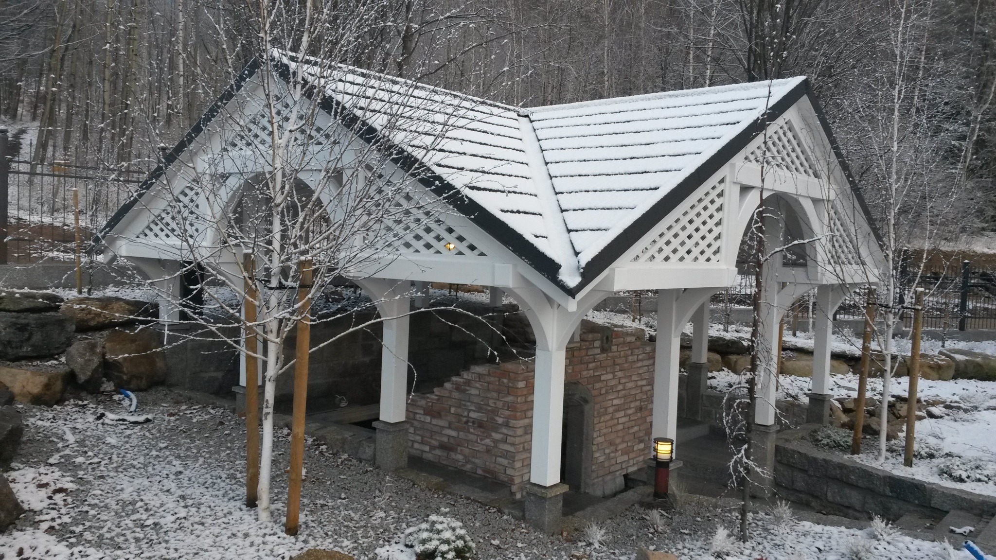 Zadaszona konstrukcja z białego drewna z kratownicą i czarnym dachem pokrytym śniegiem, wsparta na białych kolumnach z ceglaną podstawą, otoczona zimowym krajobrazem z drzewami i śniegiem.