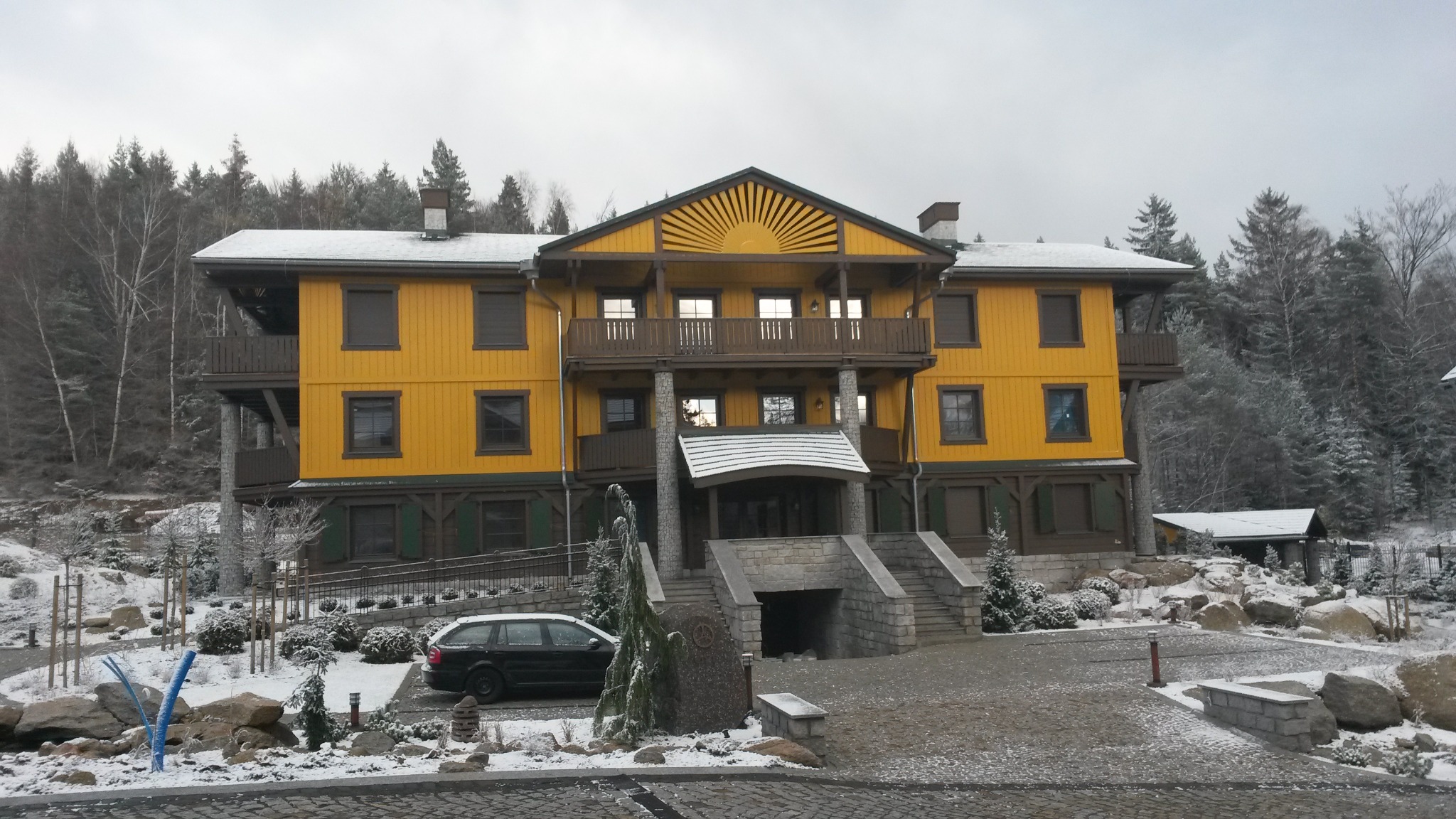 Dwukondygnacyjny budynek z żółtą elewacją i drewnianymi elementami, w zimowej scenerii z zalesionym tłem i zaparkowanym samochodem osobowym.