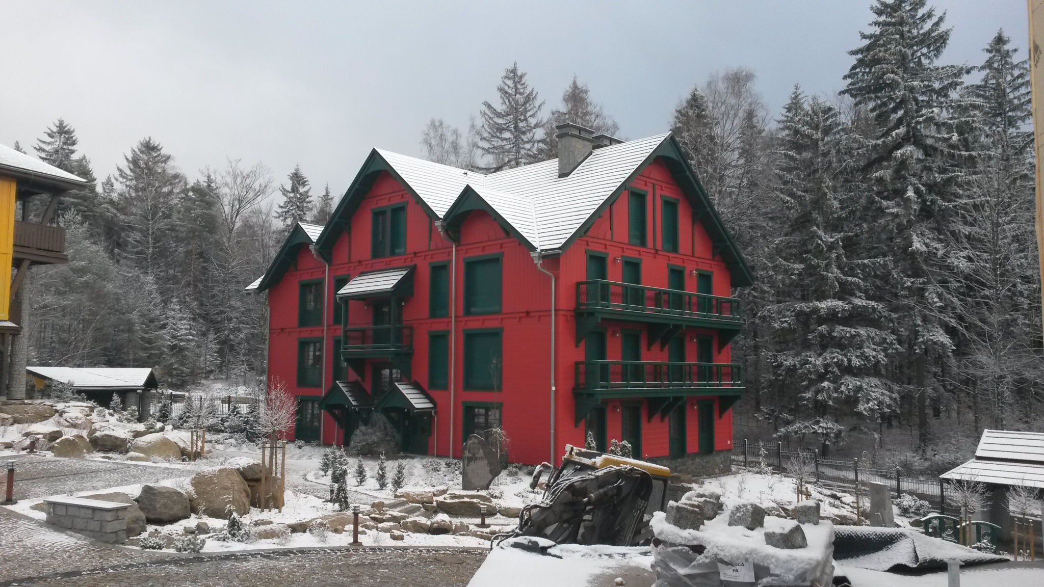 Czerwony, trzypiętrowy budynek z zielonymi akcentami i balkonami w Sidzinie, pokryty śniegiem, otoczony zimowym krajobrazem i kamieniami.