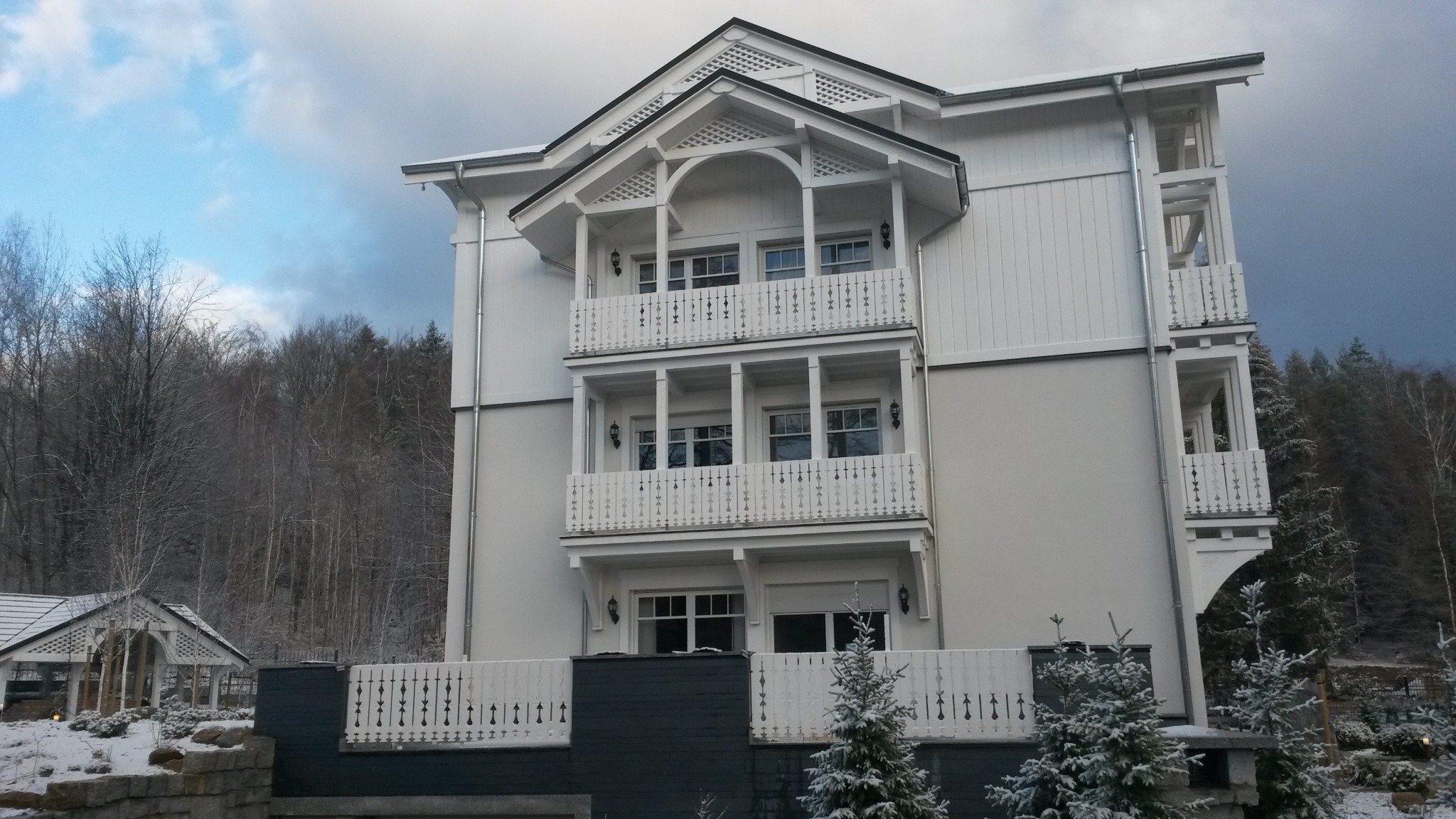 Nowoczesny, trzypiętrowy dom z białymi ścianami i balkonami z ażurowymi balustradami, otoczony zimowym krajobrazem i niewielkim budynkiem gospodarczym.