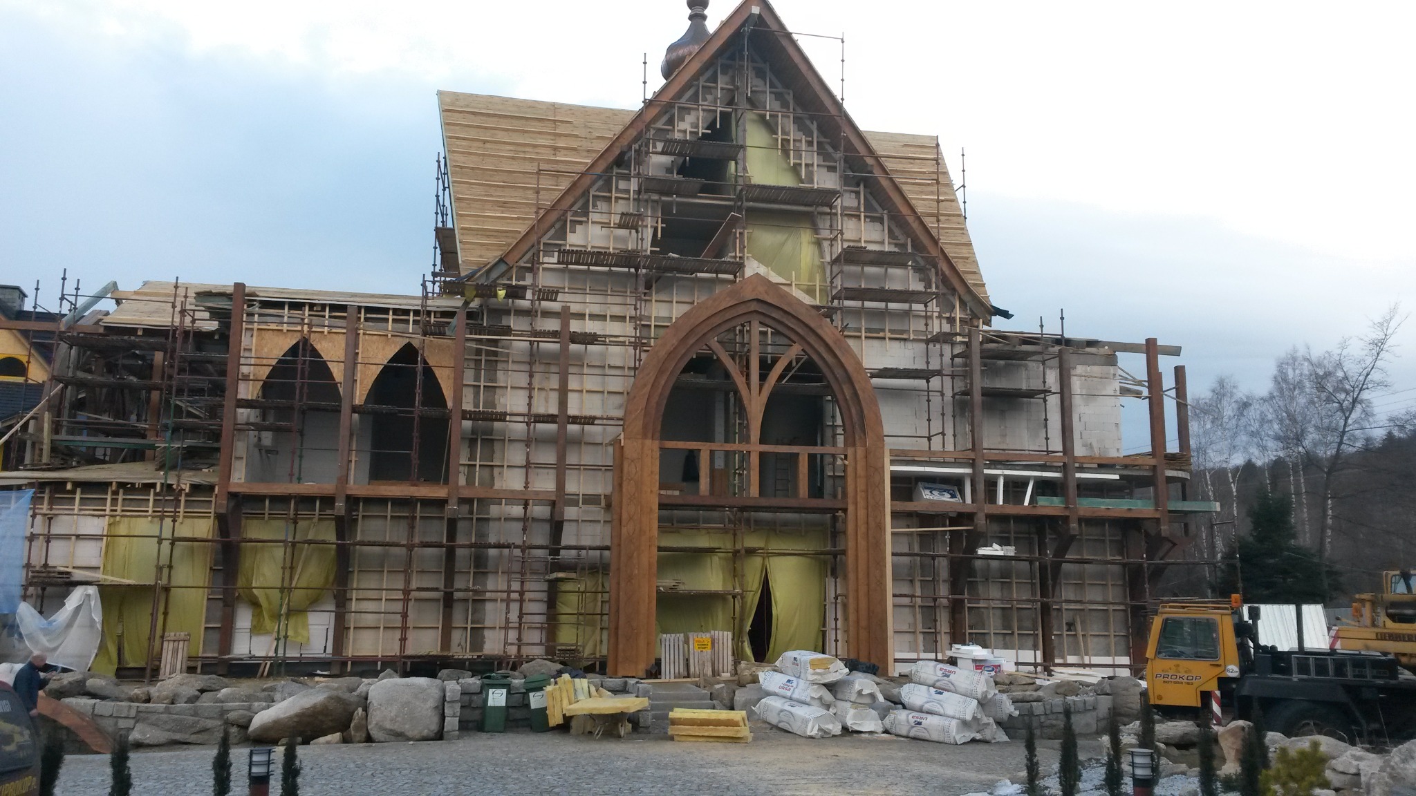 Konstrukcja budynku z drewnianym szkieletem w trakcie montażu, widoczne rusztowania, okna w stylu gotyckim i materiały budowlane na placu budowy.