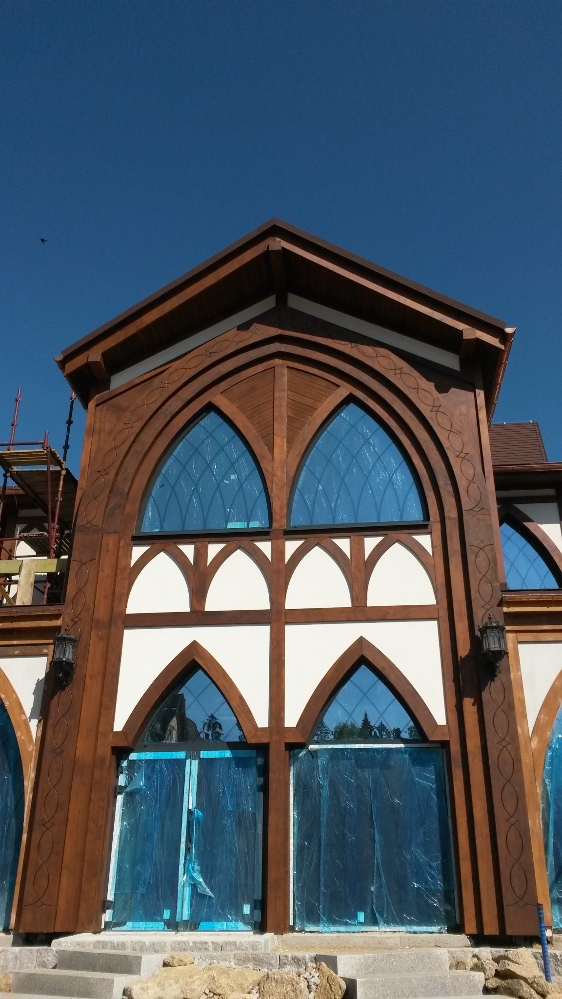 Fasada domu szkieletowego z dużymi, łukowatymi oknami zabezpieczonymi folią, widoczna dekoracyjna obróbka drewna i rusztowanie w tle.
