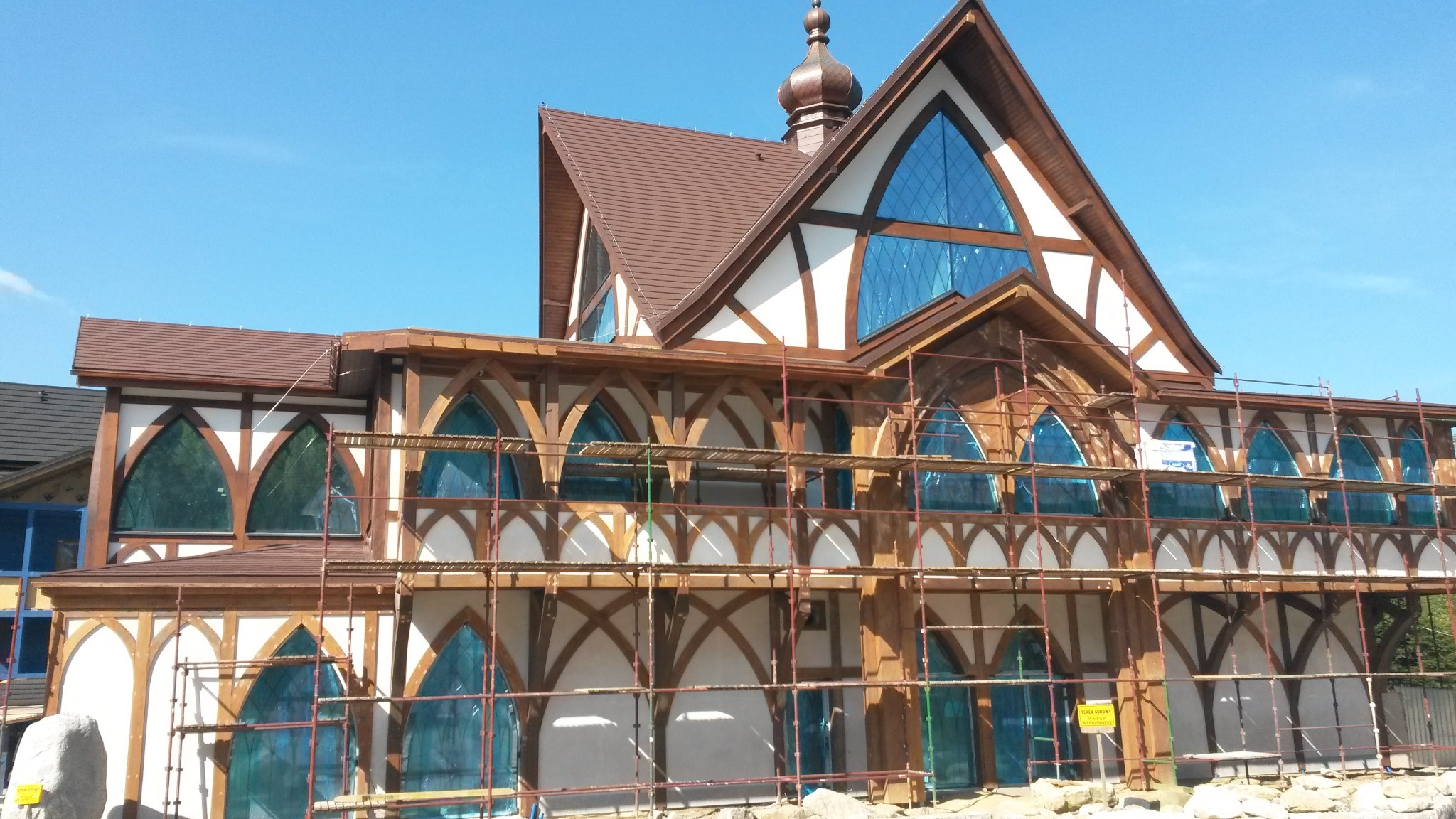 Budynek w trakcie budowy z drewnianą konstrukcją szkieletową, zabezpieczony folią i otoczony rusztowaniami, w tle błękitne niebo.