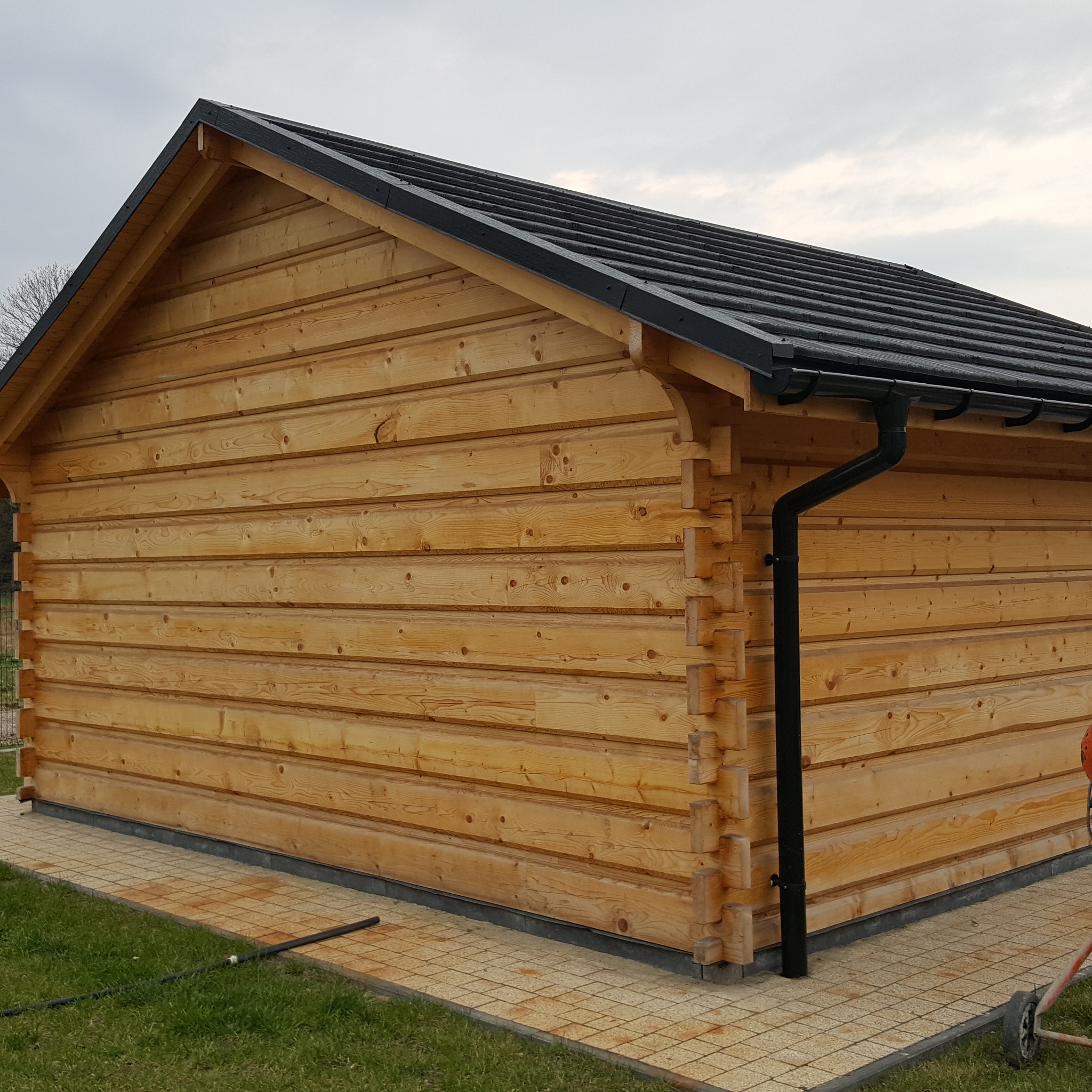 Zewnętrzny widok na mały budynek z bali o jasnym drewnie, z czarnym dachem i rynnami, osadzony na płytkach chodnikowych.