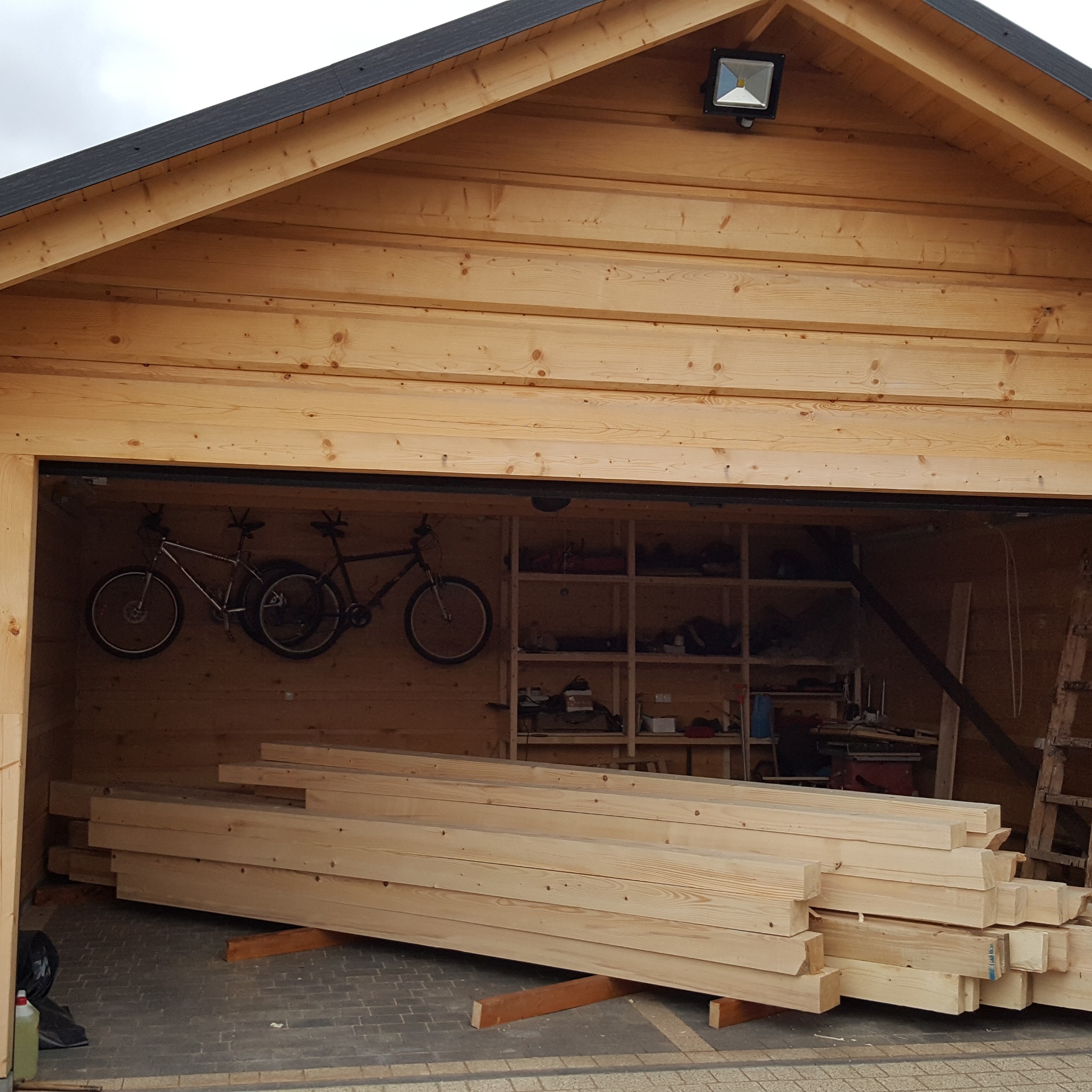 Wnętrze drewnianej konstrukcji z widocznymi belkami, rowerami zawieszonymi na ścianie, półkami z narzędziami w tle i stosem obrobionego drewna na pierwszym planie.