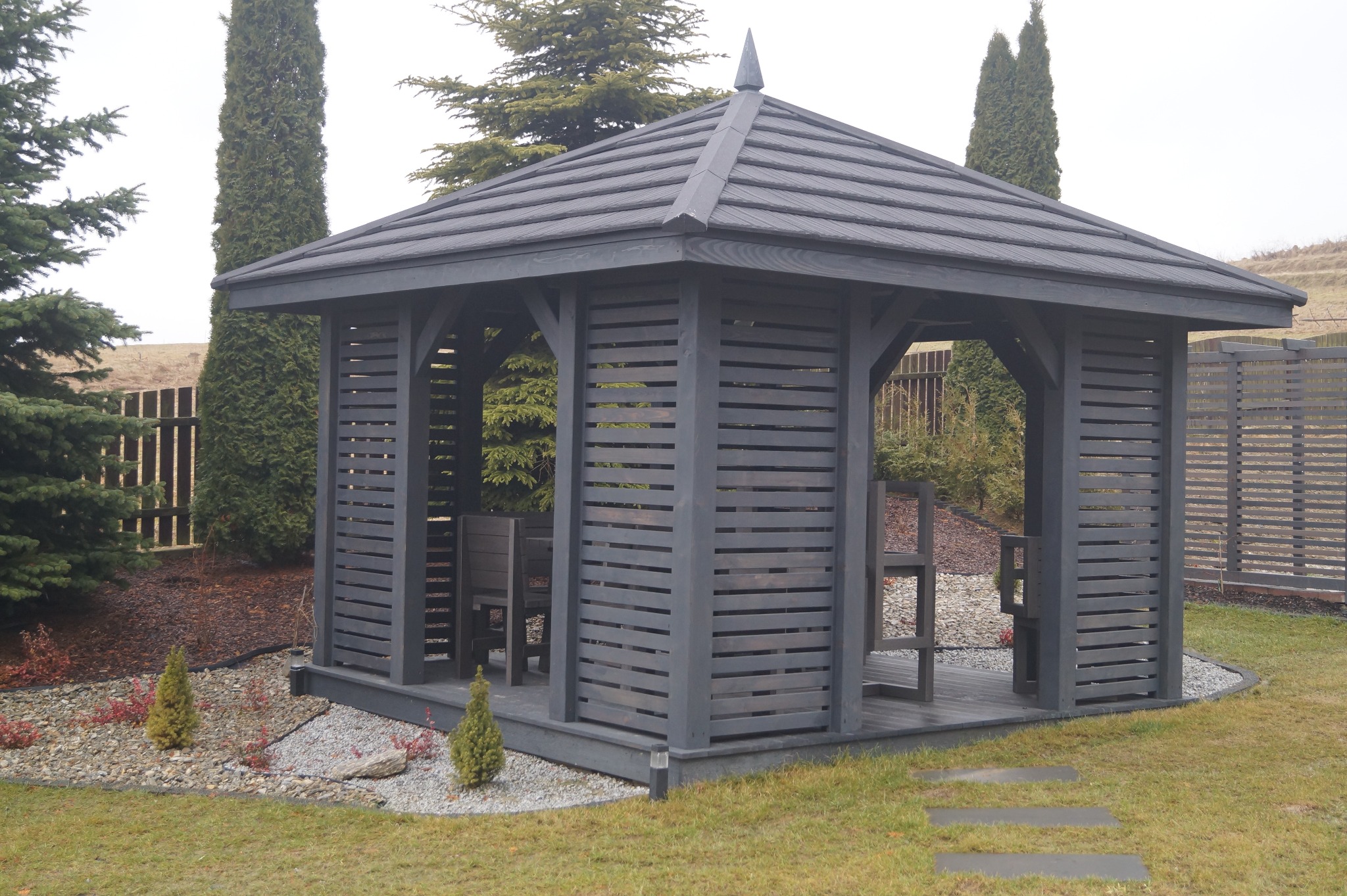 Drewniana altana pomalowana na szaro, z meblami wewnątrz, otoczona zielonym trawnikiem, ozdobnymi krzewami i kamieniami.