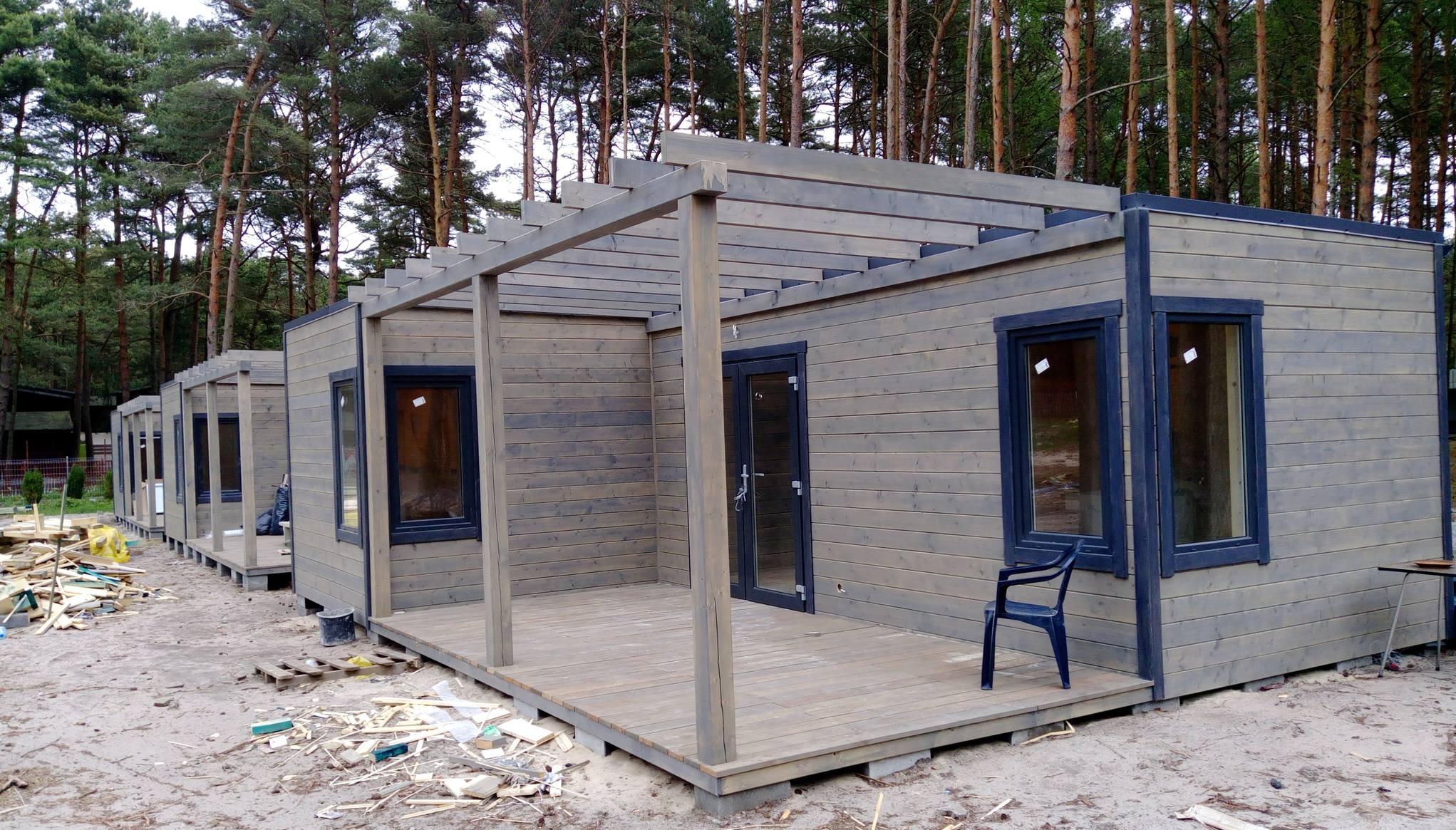 Szereg drewnianych domków letniskowych w kolorze szaro-brązowym, zadaszone drewnianą pergolą, w trakcie budowy, widoczne elementy konstrukcyjne i śmieci budowlane na piaszczystym podłożu, w tle...