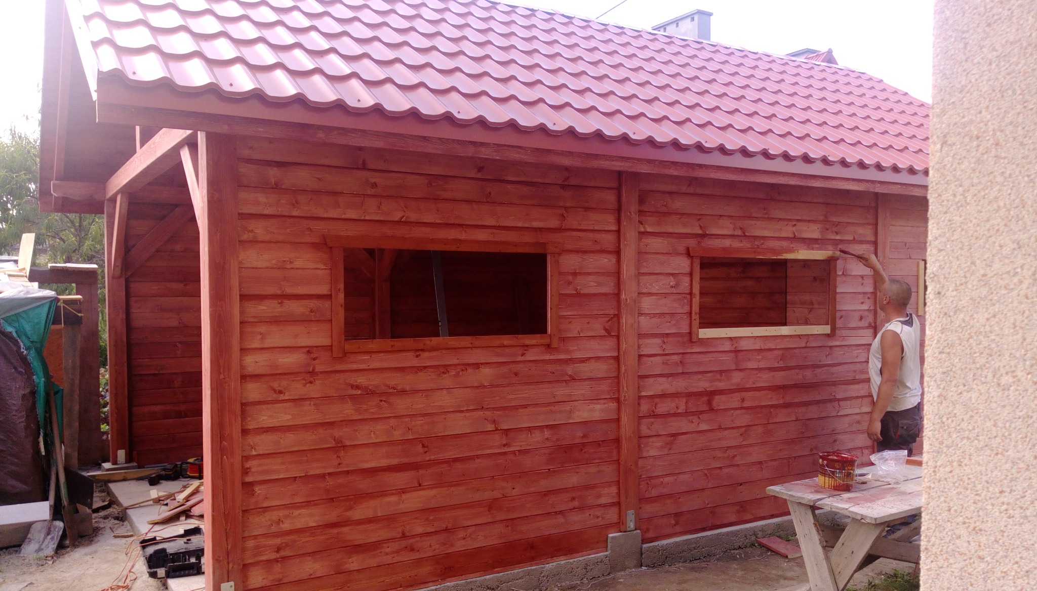 Czerwona drewniana konstrukcja w trakcie malowania, widoczne okna bez szyb, blaszany dach i mężczyzna z pędzlem.
